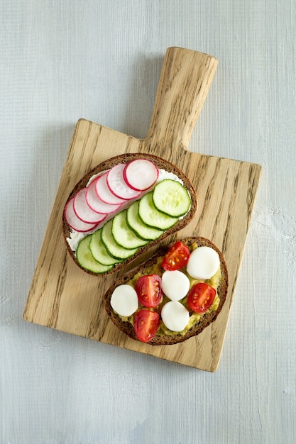 Two vegetarian sandwiches on slices of dark bread with vegetables radish, cucumber, cherry tomatoes on a wooden cutting board. Central composition on a white wooden background flat lay, top view.