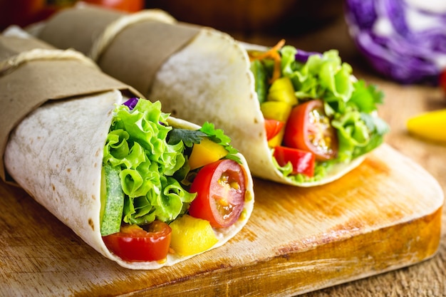 Two vegetarian tortilla wraps on wooden cutting board with vegetables in the surface