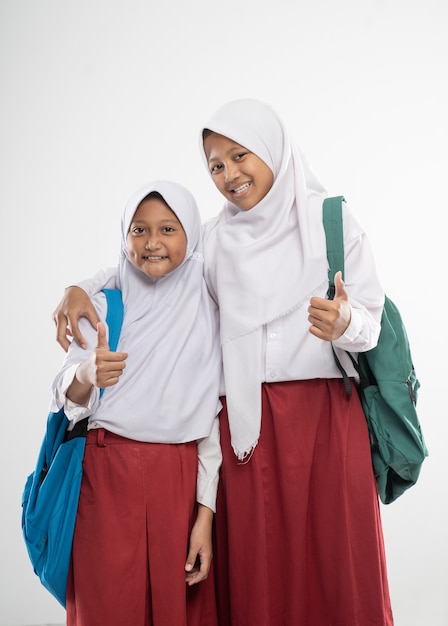 Two veiled girls in elementary school uniforms with thumbs up while carrying a backpack and a book