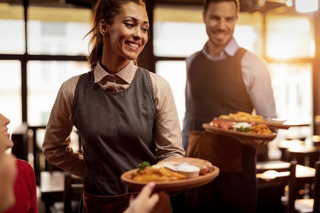 写真 居酒屋で昼食を出し、突風に食べ物を運ぶ2人のウェイターフォーカスは幸せなウェイトレスにあります