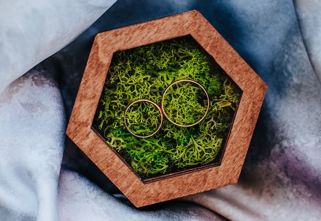 Two wedding rings in a wooden box with a plant moss on a purple fabric