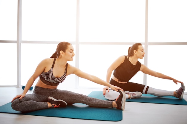 Two well-built young women stretch legs together in fitness room. Each of them reach foot with one hand. THey exercise in front of window