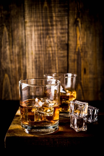 Two whiskey shot glasses on dark wooden table, with ice cubes,