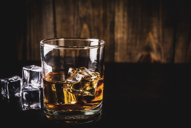 Two whiskey shot glasses on dark wooden table, with ice cubes,