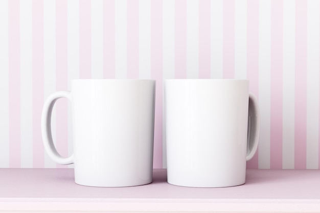 Two white coffee mugs are on a pink table with a pink striped wallpaper.