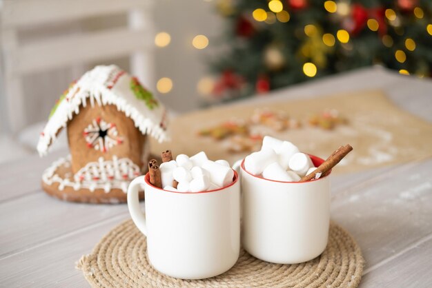 two white cup of cocoa with marshmallows and gingerbread house on table on background of Christmas t