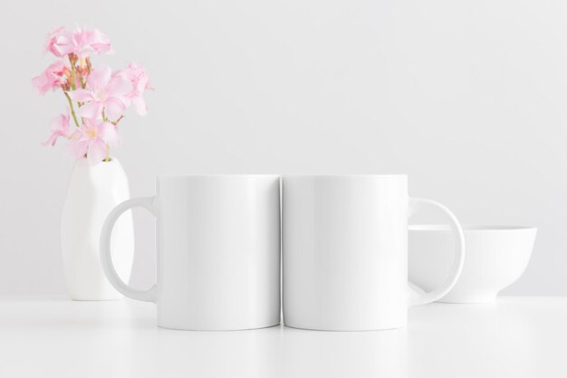 Foto mockup di due tazze bianche con un bouquet di un oleandro rosa in un vaso e una ciotola su un tavolo bianco