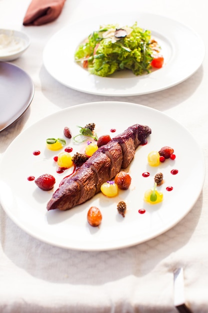 메인 코스와 샐러드가 포함된 두 개의 흰색 접시에 붉은 소스와 다양한 말린 베리와 과일을 곁들인 긴 사슴 고기