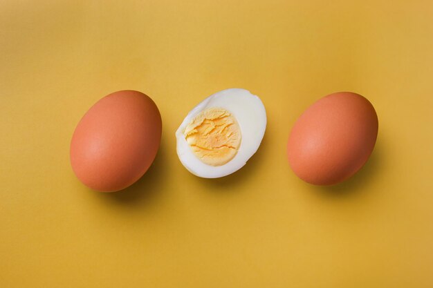 Two whole brown eggs and a boiled egg with yolk cut in half on an isolated light yellow background