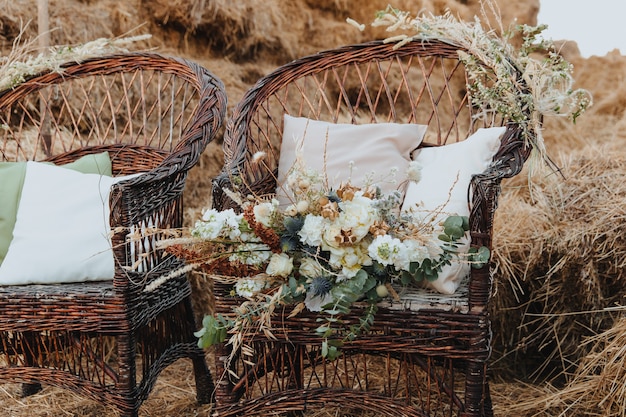 Two wicker chairs with white cushions and a bouquet of flowers on it. Rattan chairs with straw bales in the background. Boho style