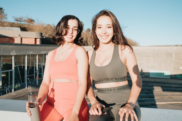 Two woman friends sitting outdoors resting after workout\
exercise dressed in sport clothes training losing weight with\
friends together smiling to camera happy african arab people doing\
exercise