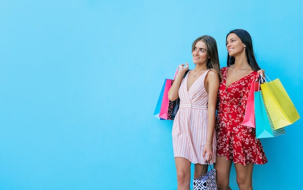 Two women are posing with their heads slightly turned towards each other, hugging, smiling with joy and showing several shopping bags in their hands