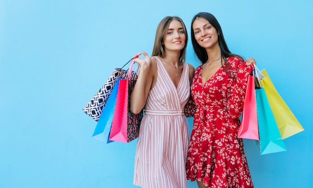 Two women are posing with their heads slightly turned towards each other, hugging, smiling with joy and showing several shopping bags in their hands