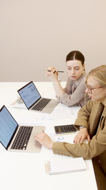 two women are sitting at a desk with laptops and a pen in the middle of them