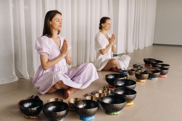 two women are sitting with tibetan bowls in the lotus position before a yoga class in the gym