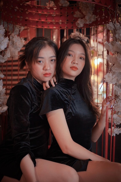 Two women in black dresses pose for a photo in front of a white flowered wall.