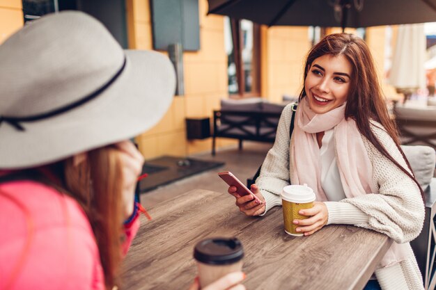 屋外カフェでコーヒーを飲みながらおしゃべりしている2人の女性。電話を使って幸せな友達。女の子が出かける