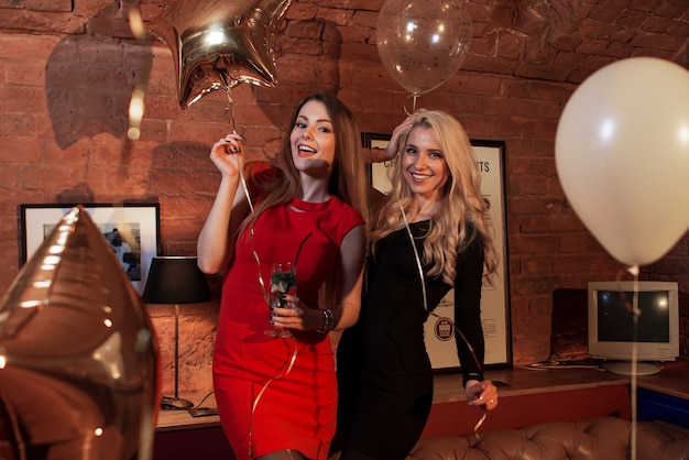 Two women in cocktail dresses posing with balloons at birthday party in stylish cafe.