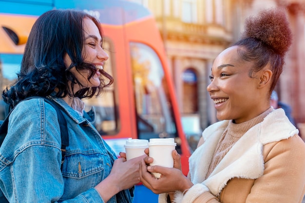 Two women in a denim jacket is talking to each other drinking coffee and waiting for a tram