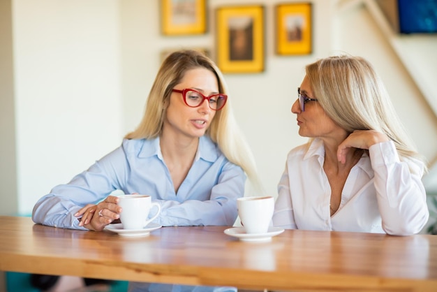 世代の異なる 2 人の女性がカフェでコーヒーを飲み、噂話をするガールフレンドがテーブルに座っておしゃべりし、カジュアルな会話をする