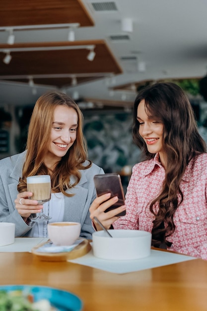 two women drinking coffee while sitting in cafe or restaurant and looking on mobile phone