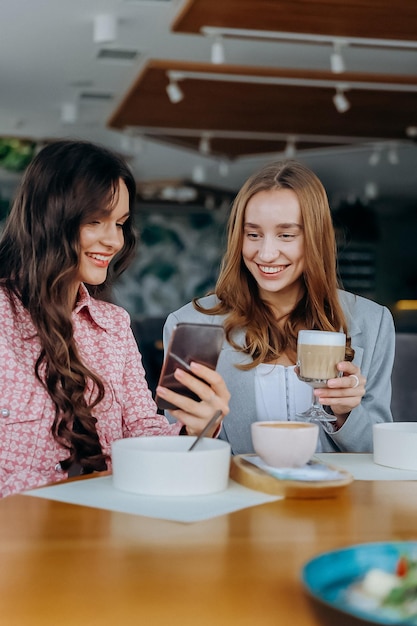two women drinking coffee while sitting in cafe or restaurant and looking on mobile phone