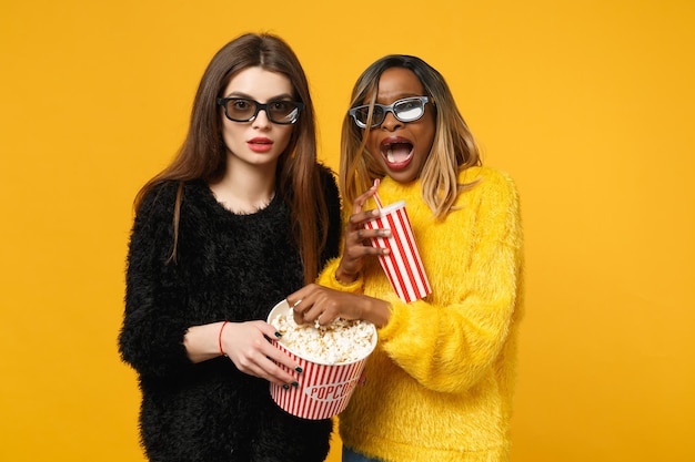 Two women friends european and african american in black yellow clothes hold bucket of popcorn isolated on bright orange wall background, studio portrait. People lifestyle concept. Mock up copy space.