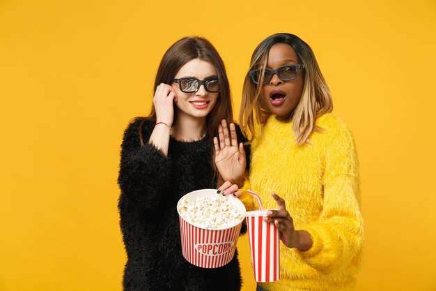 Two women friends european and african american in black yellow clothes hold bucket of popcorn isolated on bright orange wall background, studio portrait. People lifestyle concept. Mock up copy space.