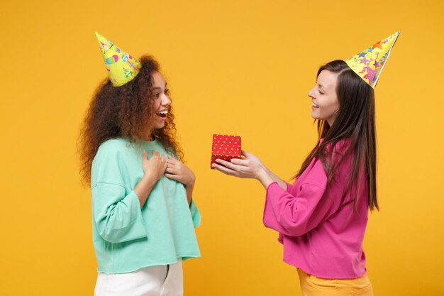 Two women friends european african american girl in pink green clothes birthday hats posing isolated on yellow background. People lifestyle concept. Mock up copy space. Hold red present box with gift.