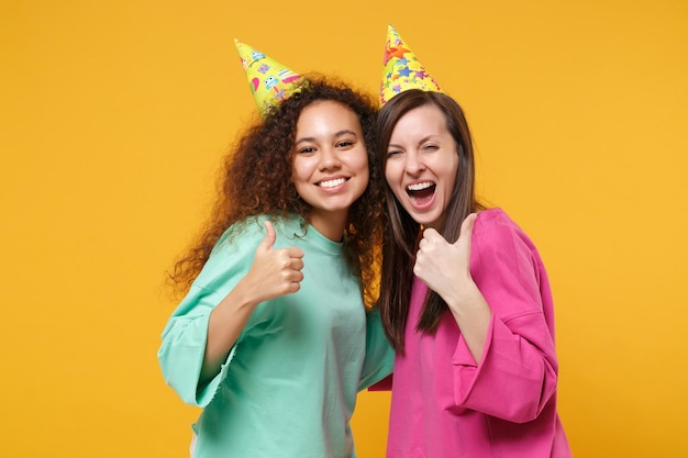 Two women friends european and african american girls in pink green clothes birthday hats posing isolated on yellow background. People lifestyle concept. Mock up copy space. Hugging showing thumbs up.