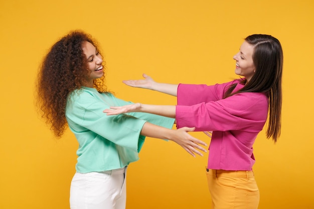 Two women friends european and african american girls in pink green clothes posing isolated on yellow background. People lifestyle concept. Mock up copy space. Stand with outstretched hands for hugs.