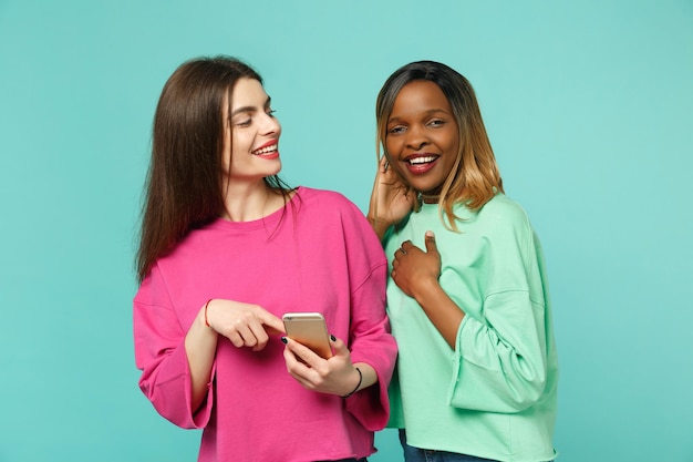 Two women friends european and african american in pink green clothes hold in hand cellphone isolated on blue turquoise wall background, studio portrait. People lifestyle concept. Mock up copy space.