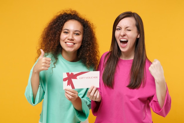 Two women friends european african girl in pink green clothes\
posing isolated on yellow background. people lifestyle concept.\
mock up copy space. hold gift certificate showing thumb up winner\
gesture.