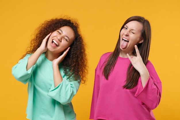 Two women friends european african girls in pink green clothes posing isolated on yellow background. people lifestyle concept. mock up copy space. cover ears with hands, showing heavy metal rock sign