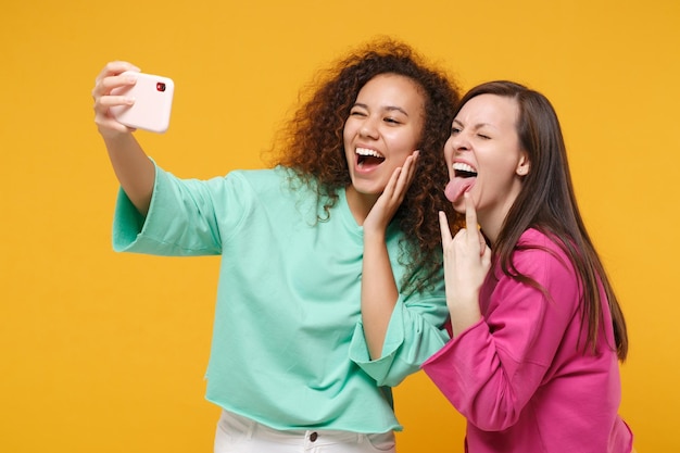 Two women friends european american girls in pink green clothes posing isolated on yellow background. people lifestyle concept. mock up copy space. doing selfie shot on mobile phone, showing horns up