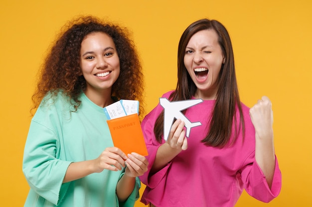 Photo two women friends european american girls in pink green clothes posing isolated on yellow background. people lifestyle concept. mock up copy space. hold passport ticket airplane doing winner gesture.