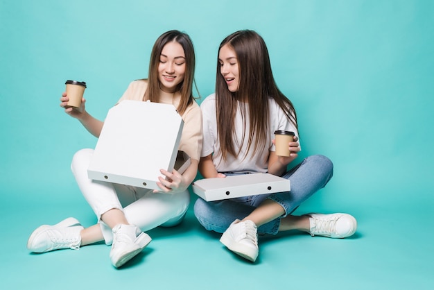 Two women friends sitting on the floor drink coffee to go and eat pizza isolated on turquoise wall