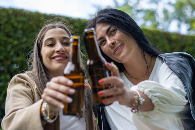 Two women friends sitting on the grass toasting with bottles of beer Lifestyle of young friends in hats sitting on the grass toasting with wine