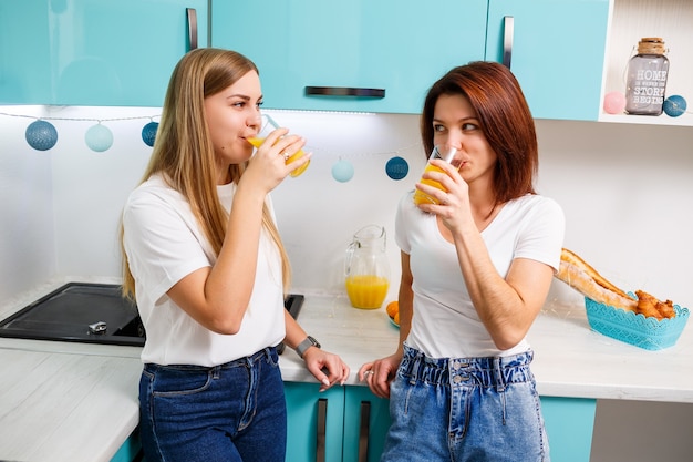 Two women friends standing in the kitchen and drinking orange juice. Girlfriends chat and share secrets in the kitchen, breakfast