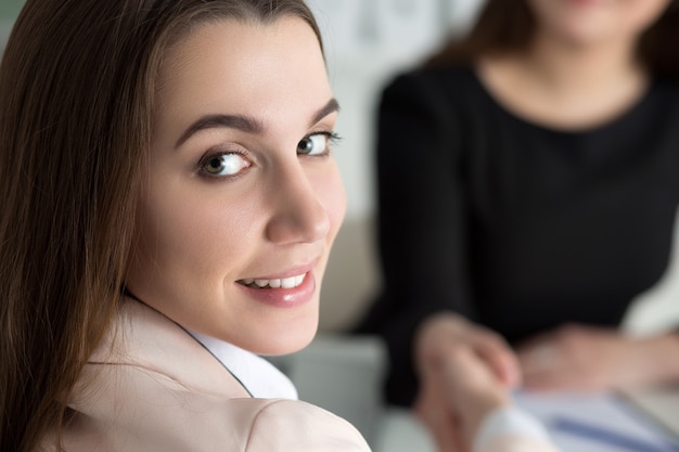 Two women handshake in office. Businesswomen shaking hands. Lady looks back over her shoulder. Partnership, business and collaboration concept. Partners made deal. Formal greeting gesture