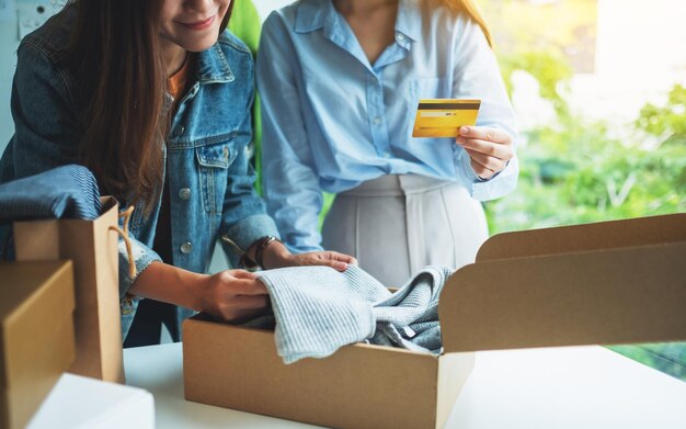 Two women holding and using credit card for online shopping while opening a postal parcel box of clothing at home