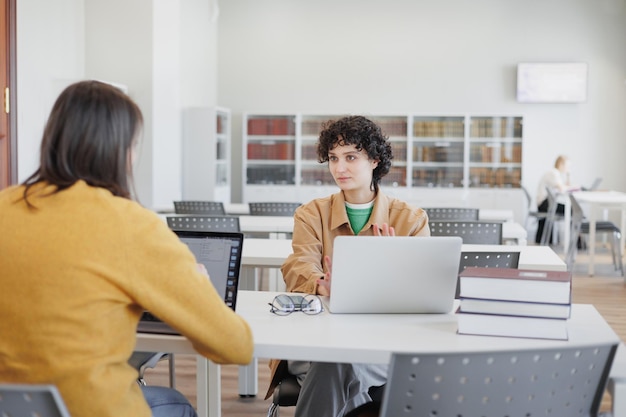 Two women in library or coworking are sitting opposite each other working on laptop