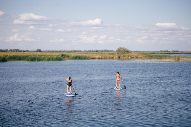 Two women of middle age sup boarding in middle of small blue lake covered with ripples with puddles looking at one direction wearing swimming suits Active lifestyle for older people