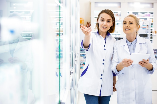 Two women pharmacists studying a shop window in a pharmacy discussing a range of medicines