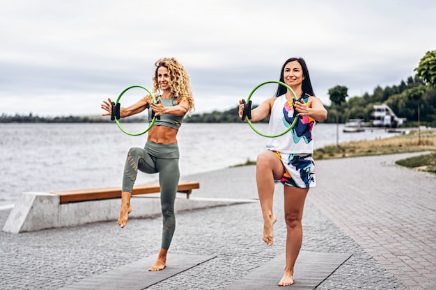 Two women practice yoga exercises on a mat with a special sports circle, on the street near the water. Active lifestyle. Yoga concept