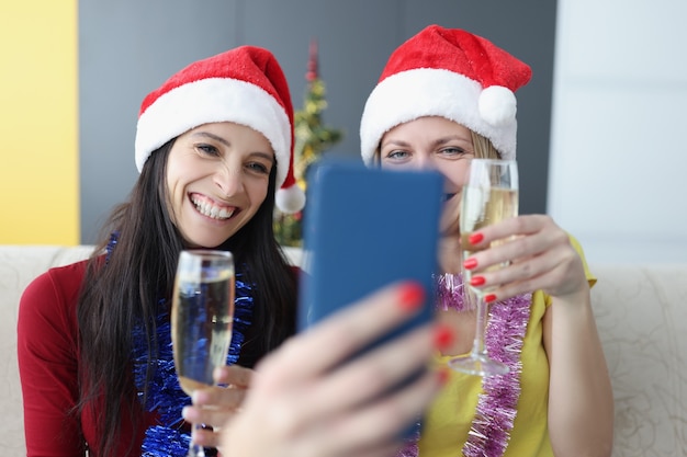 Two women in santa hats drinking champagne and taking selfie on cell phone at home. Removed merry christmas and happy new year greetings concept