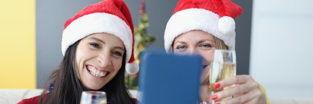 Two women in santa hats drinking champagne and taking selfie on cell phone at home