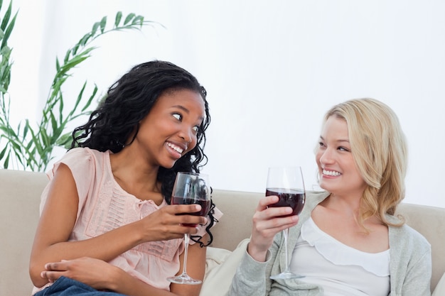 Two women sitting on a couch are holding wine glasses 