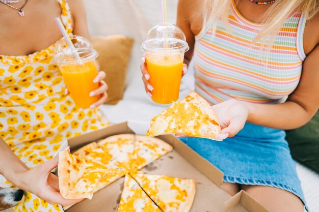 Two women sitting on a sofa in the backyard and eating pizza, drinking lemonade on summer sunny day