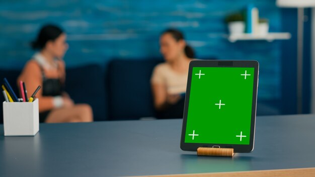 Two women sitting on sofa talking about digital technology. Office room is equipped with isolated tablet computer with mock up green screen chroma key display standing on table desk